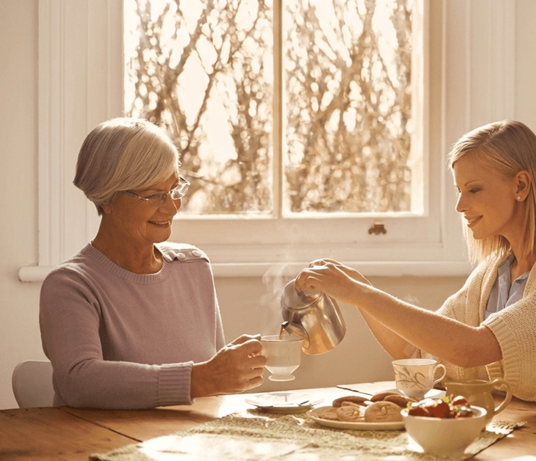 Two generations sharing a moment—parent and adult child at a table with tea, warm and connected
